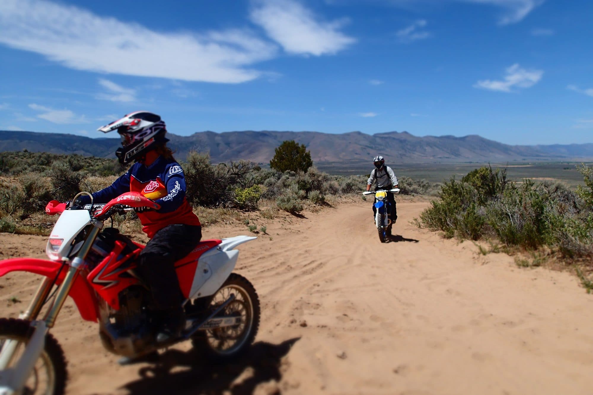 Two riders on dirt bikes on a sandy off-road trail through sagebrush hills.