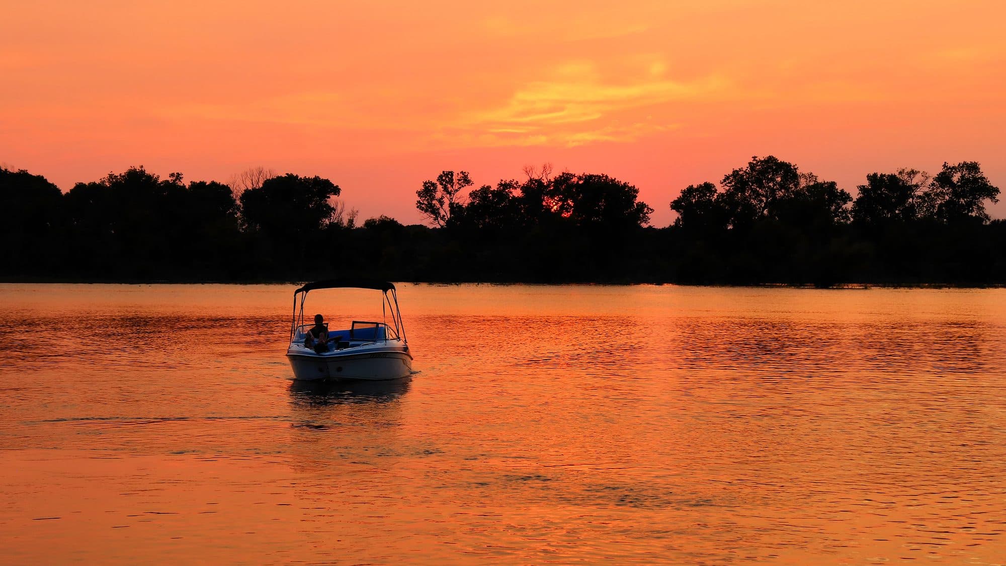 A small motorboat on Lake Whitney, Texas, at sunset, with the water and sky glowing orange behind a line of dark trees.