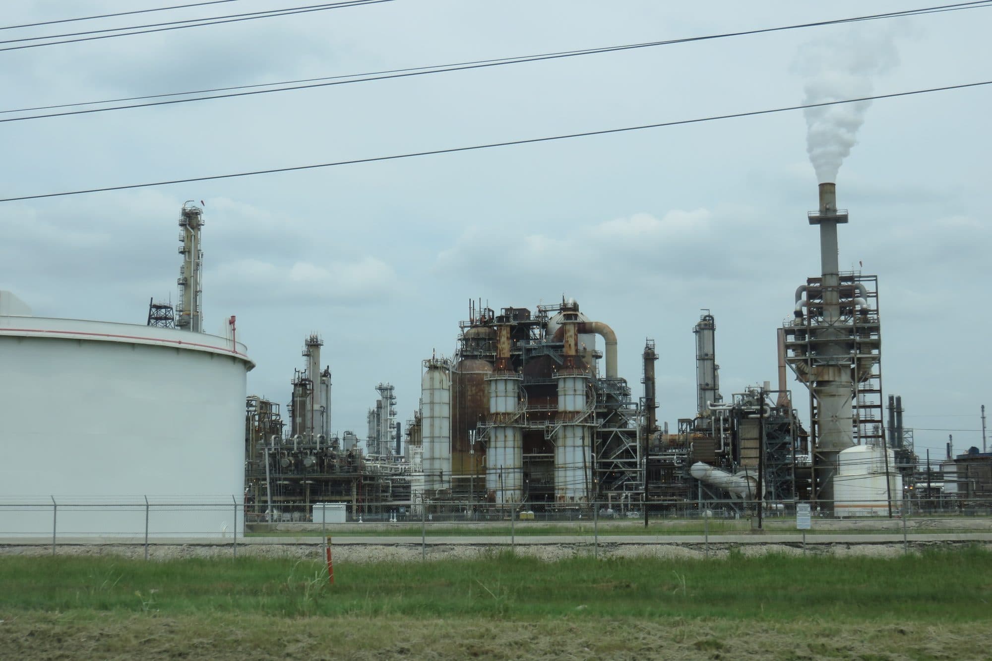 The towers and pipes of an oil refinery in Houston, with steam rising from a stack behind a white storage tank.
