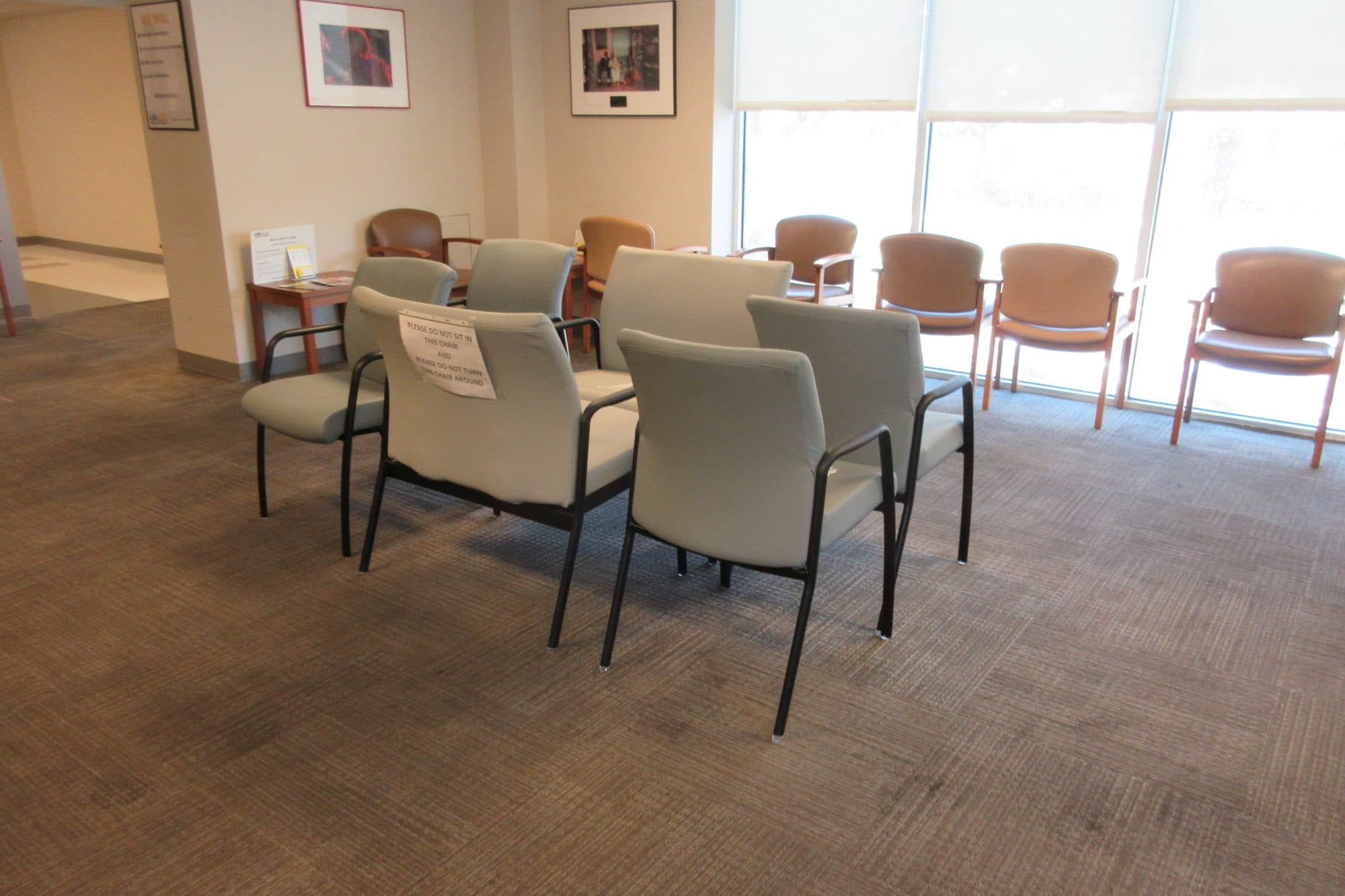 An empty, sunlit waiting room with padded chairs spaced apart on a carpeted floor.