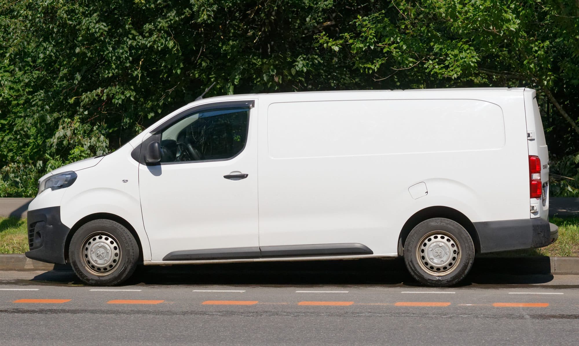A plain white cargo van parked along a street next to green trees.