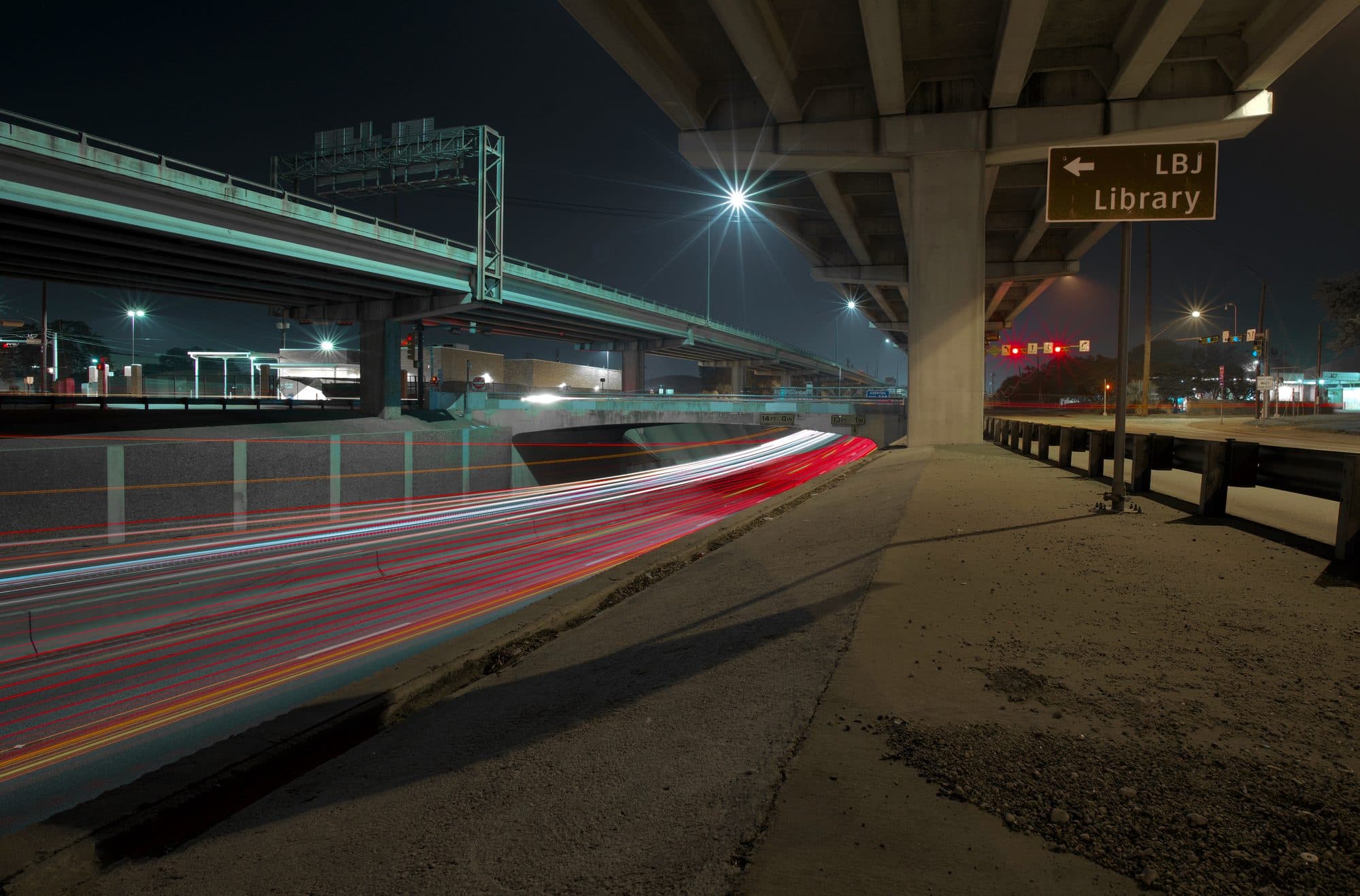 Red and white light trails from traffic passing beside a freeway overpass in Austin at night.