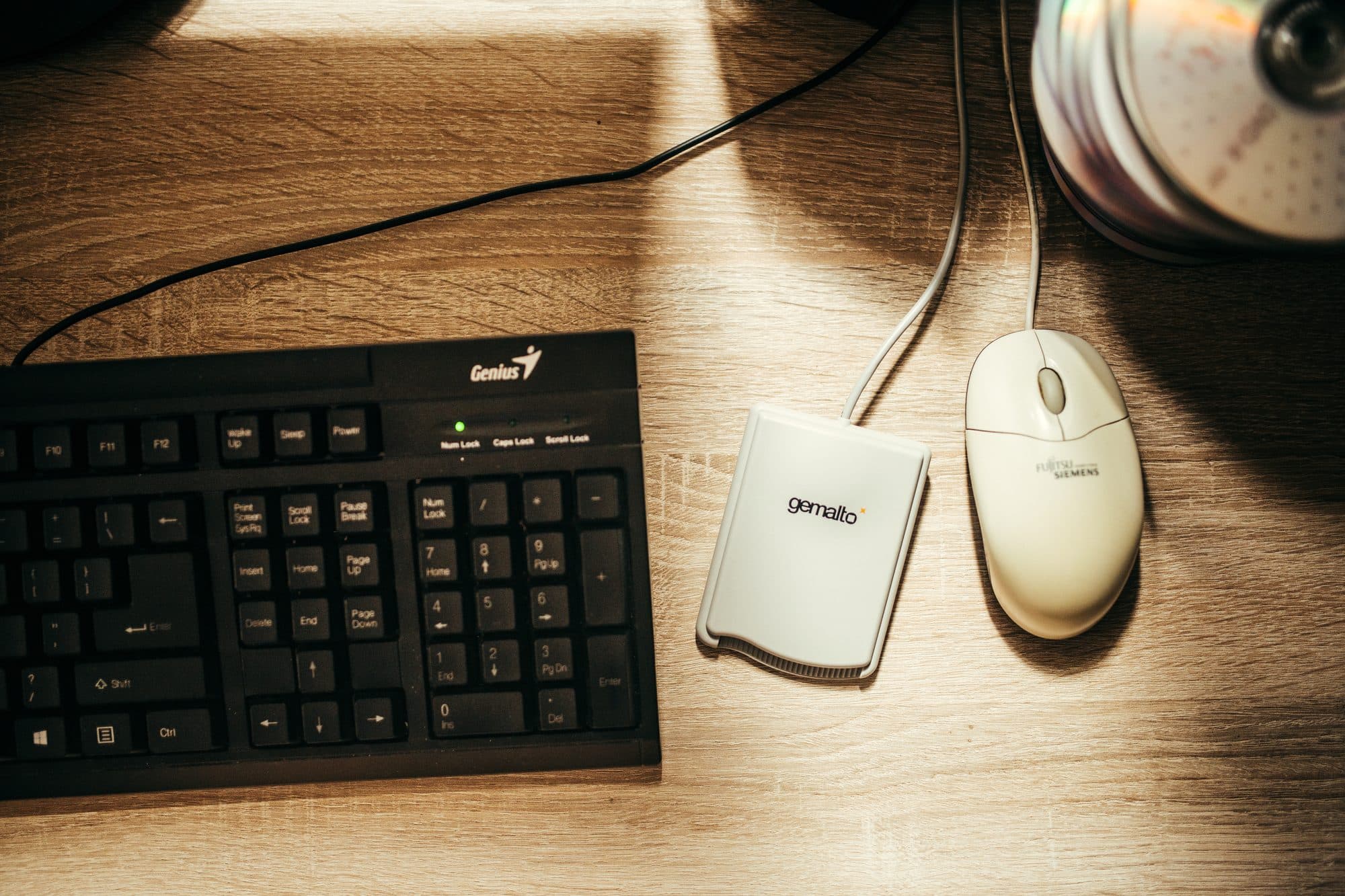 A black computer keyboard, a smart card reader and a mouse on a wooden desk lit by a lamp.