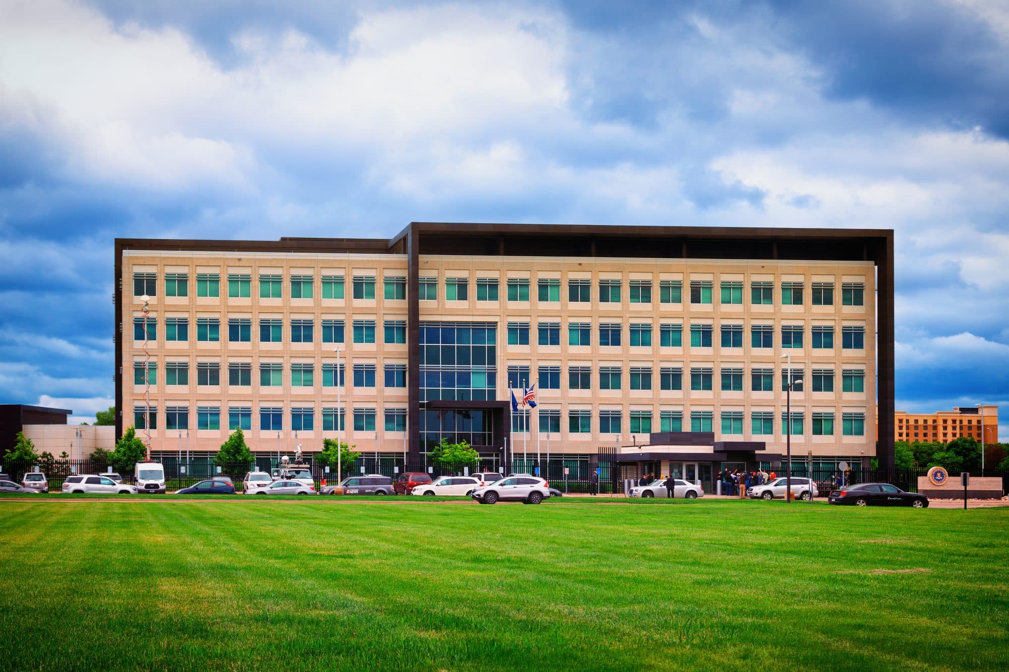 The modern FBI field office building in Minneapolis, behind a wide green lawn under a cloudy sky.