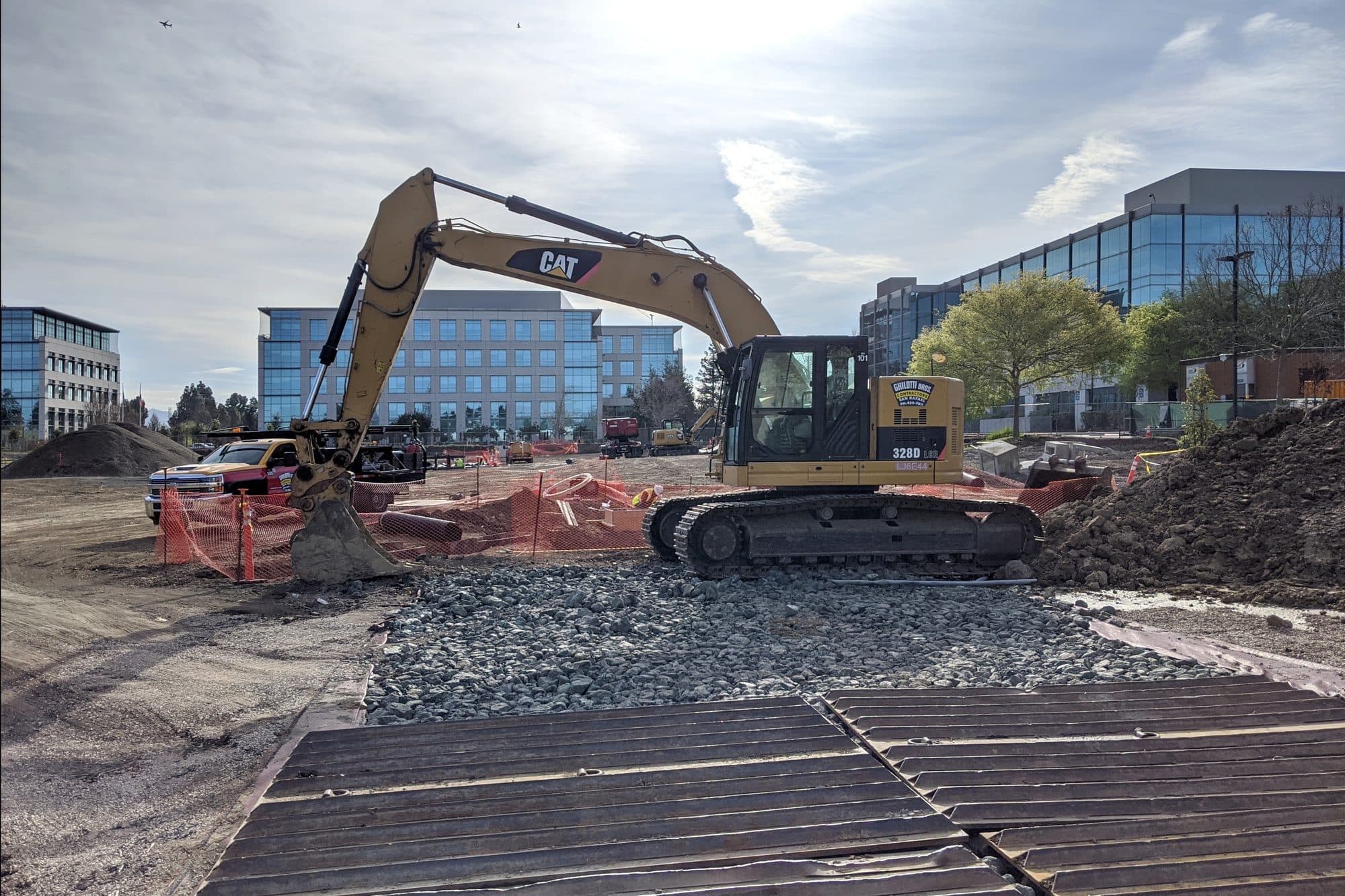 A tracked excavator working on a construction site next to office buildings.