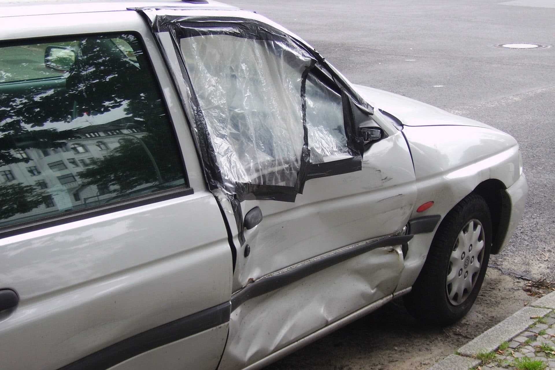 A silver car with a dented driver's door and a broken window taped over with plastic, parked by a curb.