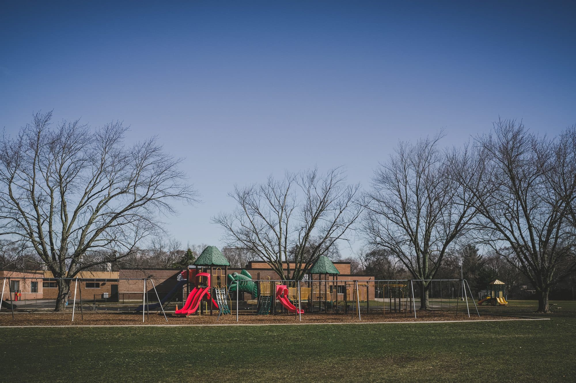 An empty playground at the edge of a grassy park under bare winter trees.