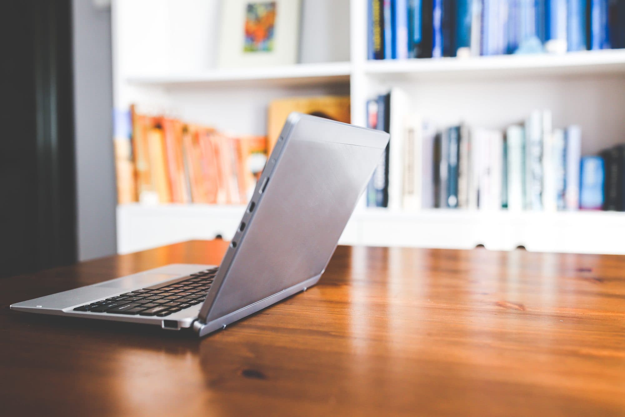 An open silver laptop on a wooden desk in front of a blurred bookshelf.