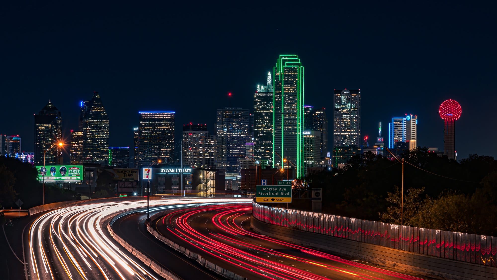 Long-exposure light trails on a curving Dallas freeway at night, with the lit downtown skyline and Reunion Tower behind.