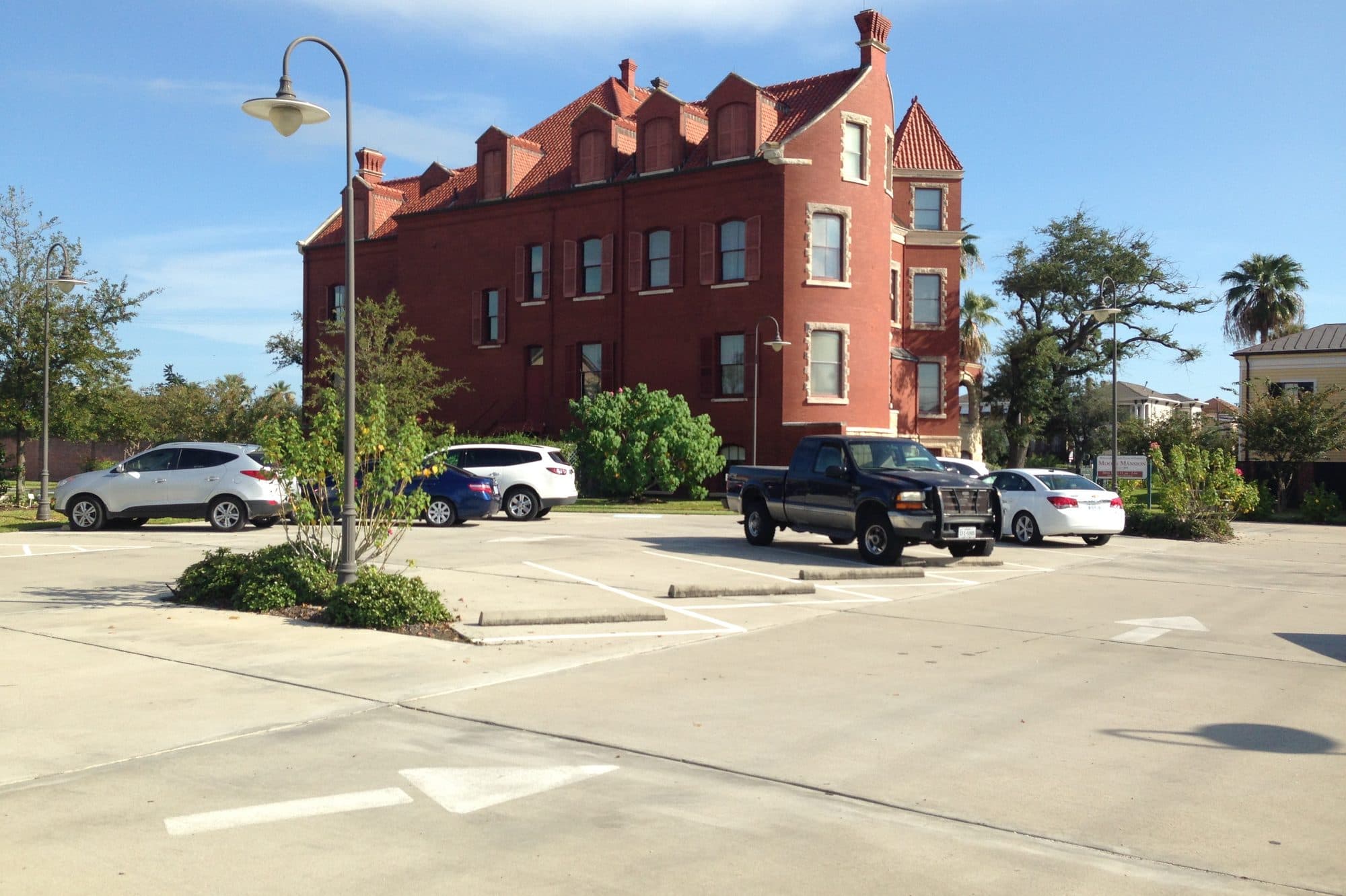 A sunny parking lot with a few cars and pickups beside the red brick Moody Mansion in Galveston, Texas.