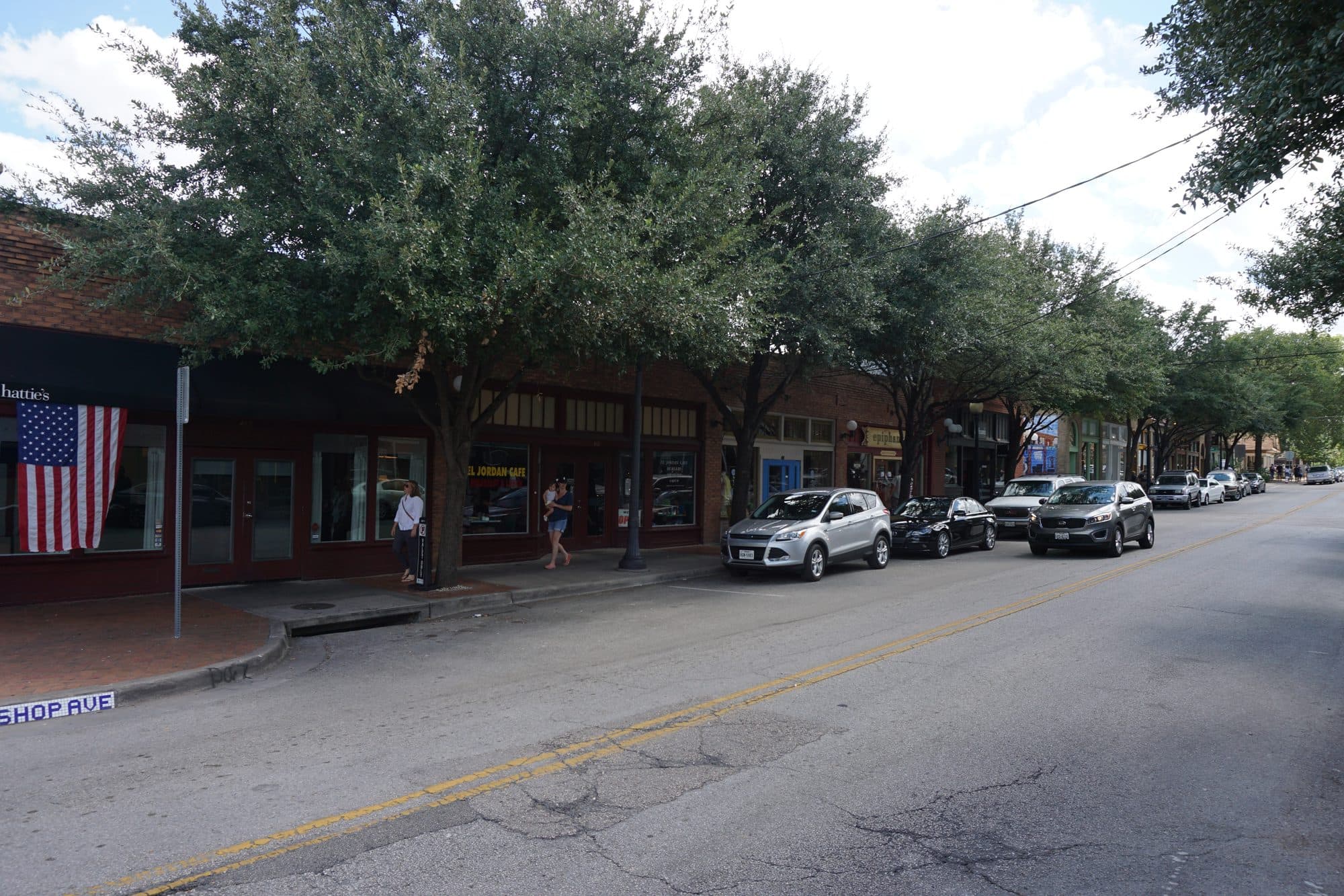 A tree-lined stretch of Bishop Avenue in Oak Cliff, with people walking on the sidewalk past parked cars.