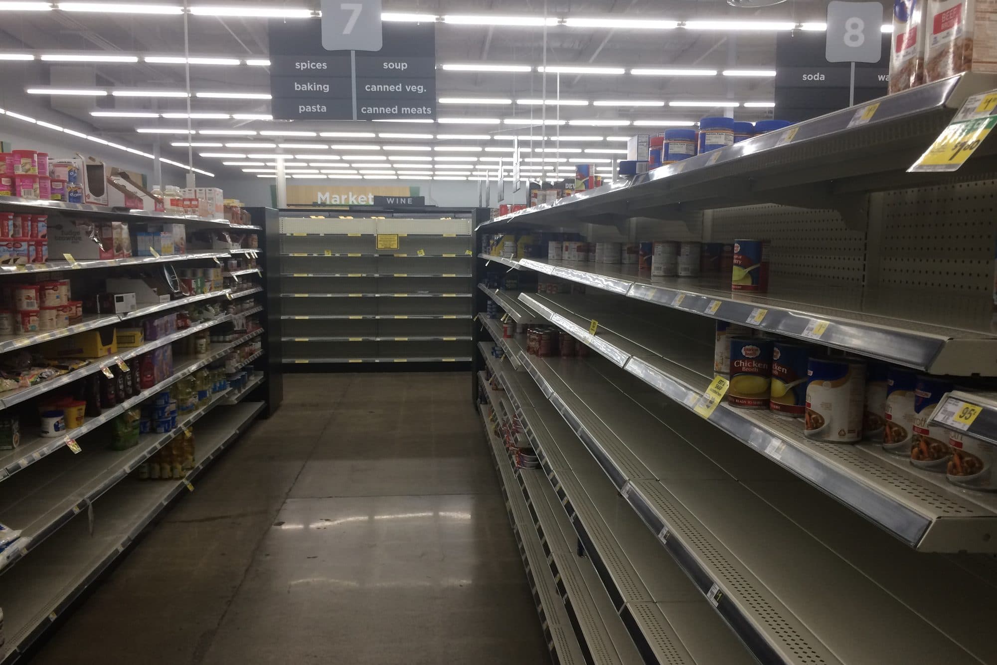 A long, quiet store aisle with half-empty shelves under bright ceiling lights.