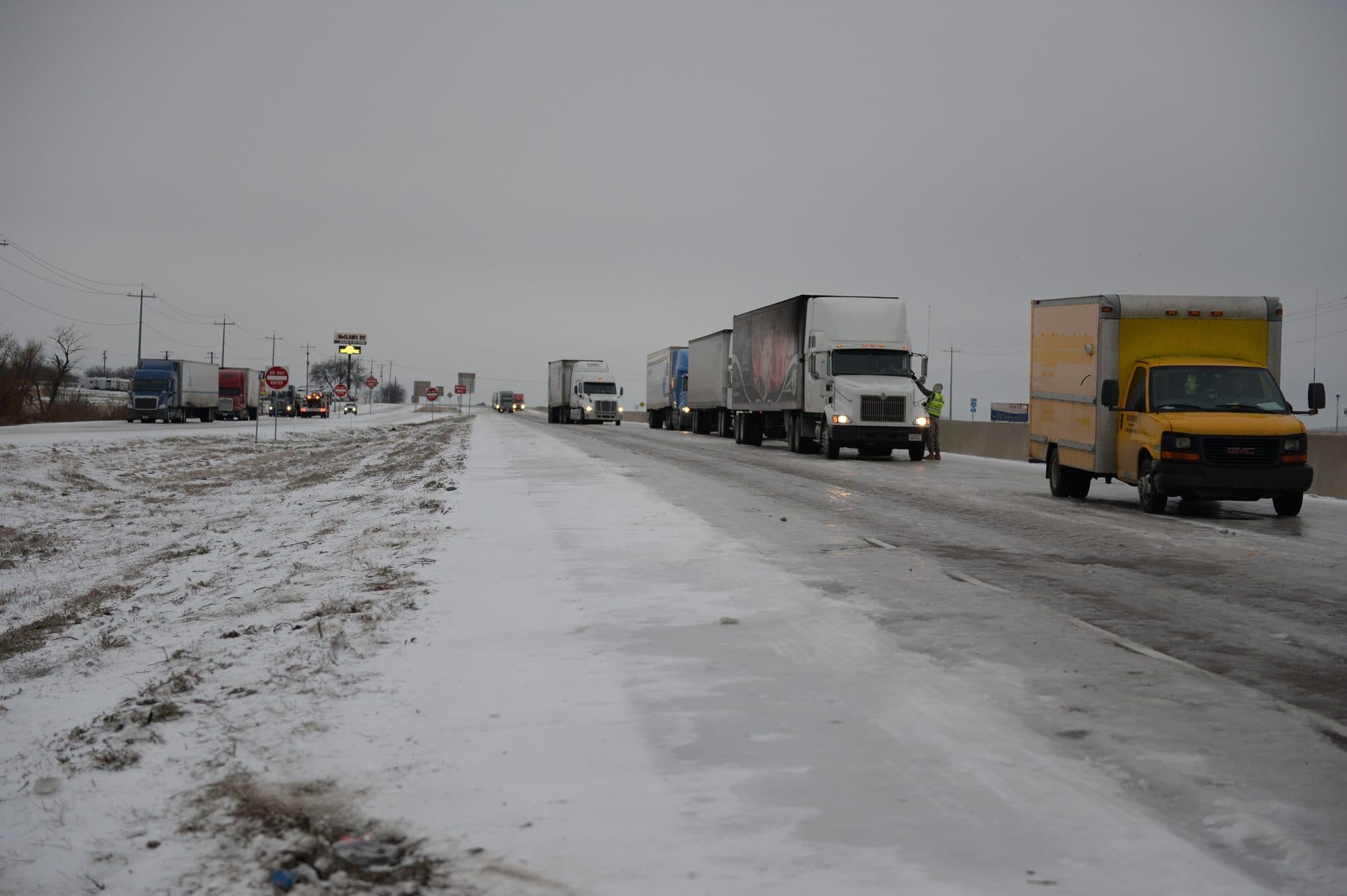 A line of 18 wheelers and a box truck on an icy Texas interstate during a winter storm.