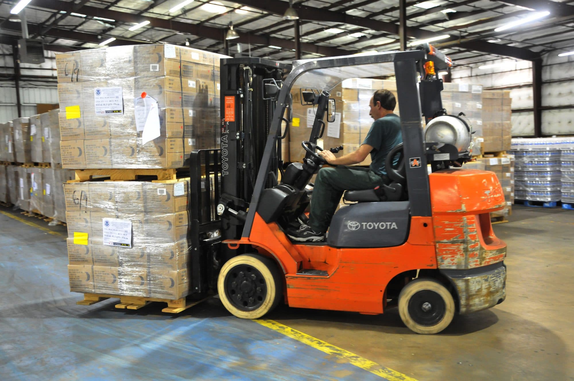 A worker driving an orange forklift loaded with boxes across a warehouse floor.